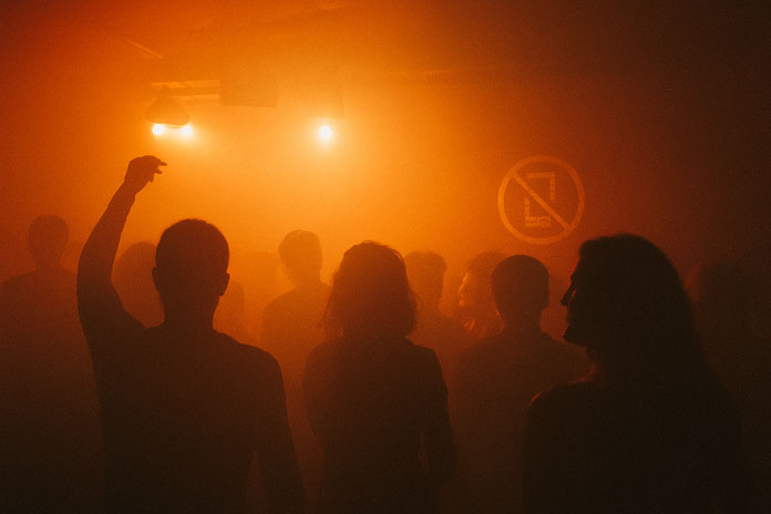 Siluete plesača u toplom crvenom svetlu; diskretan znak „no phone” na zidu.